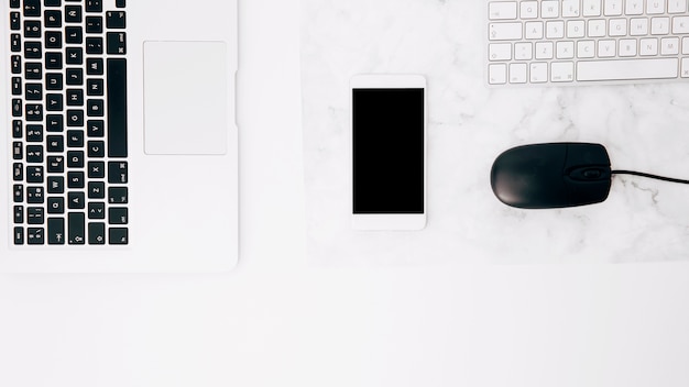 An overhead view of laptop; cellphone; mouse and keyboard on white desk Free Photo