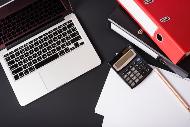 An overhead view of laptop; file folders; calculator; pencils and paper on black background Free Photo
