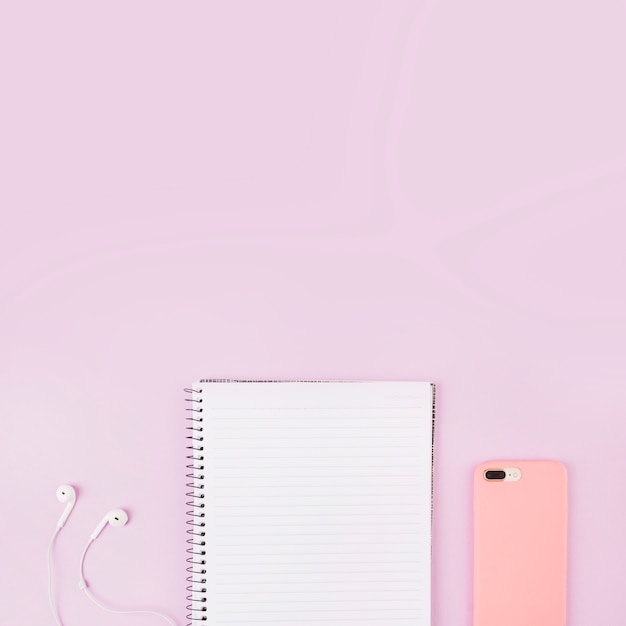 Overhead view of smartphone; earphone and notepad at the edge of pink background Free Photo