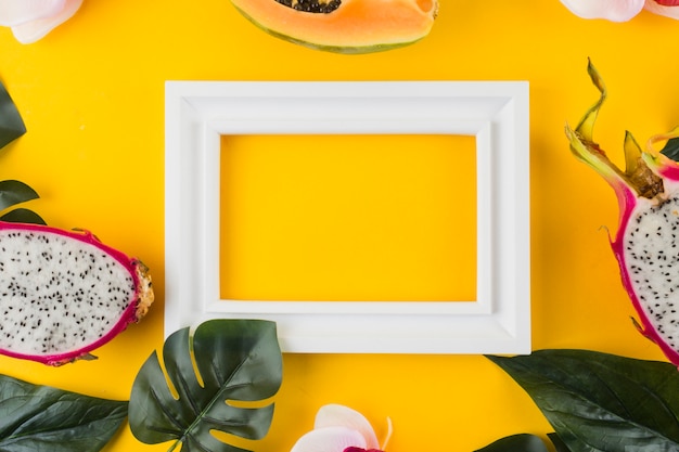 Papaya; dragon fruit; leaves around the empty white border frame against yellow backdrop Free Photo