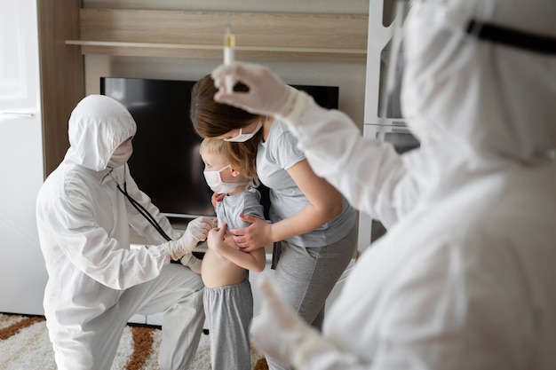 フェイスマスクと防護服で病気の子供を調べる小児科医医師 自宅で母親と一緒に病気の男の子 子供の家庭でのウイルス治療 コロナウイルスパンデミック Covid 19アウトブレイク プレミアム写真