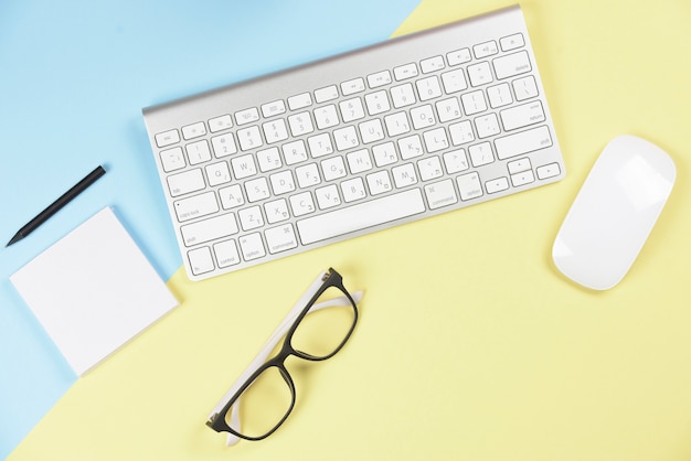Pencil; adhesive notepad; eyeglasses; wireless keyboard and mouse on blue and yellow backdrop Free Photo