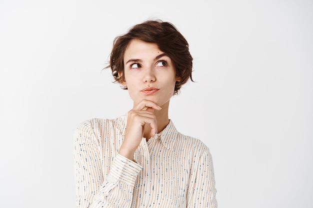Pensive girl in blouse touching chin, looking at upper right corner and thinking, making choice, standing over white wall Free Photo