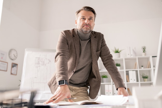 Premium Photo | Pensive mature architect leaning against desk with ...