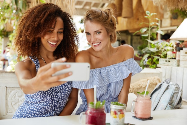 人 レジャー レクリエーションの時間 陽気なアフリカ系アメリカ人女性と彼女の親友はカフェで自由な時間を過ごし 携帯電話で自撮りを作って スムージーを飲みます 多民族の関係の概念 無料の写真