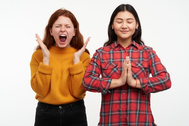 People and lifestyle concept. girl screams in anger while her friend calm meditating, fold hands in namaste sign. wearing yellow sweater and checkered shirt. stand isolated over white wall Free Photo