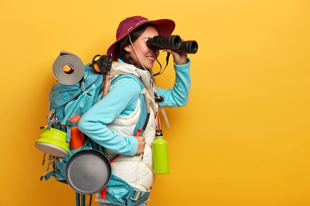 People, lifestyle, holiday, tourism concept. cheerful female tourist observes something in binoculars, stands with backpack, wears casual active wear Free Photo