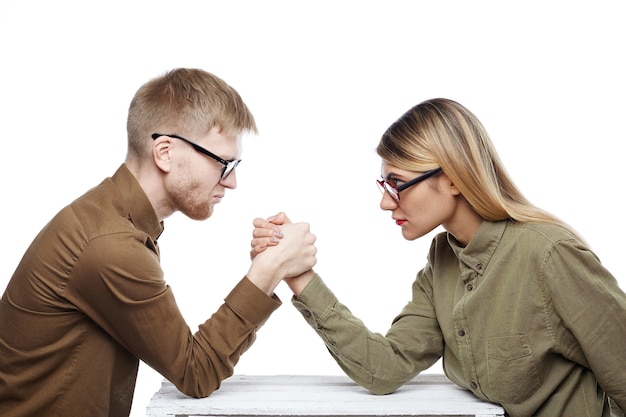 People, teamwork, cooperation and competition concept. side view of young female and bearded male colleagues both wearing glasses arm wrestling, staring at each other with confident determined looks Free Photo