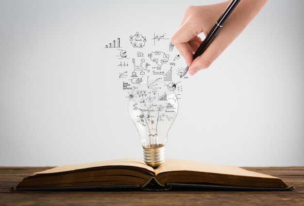 Person drawing symbols coming out of a light bulb on top of a book Free Photo