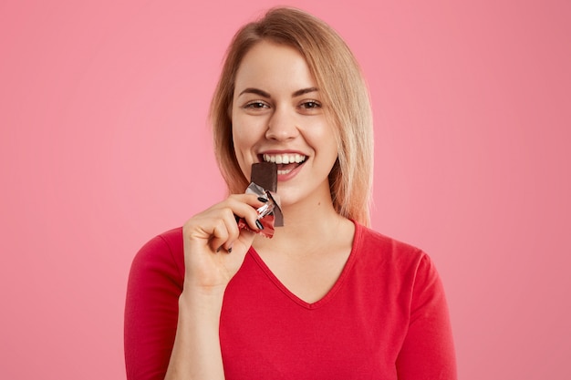Photo of attractive light haired european young woman eats delicious sweet chocolate, being sweettooth Free Photo