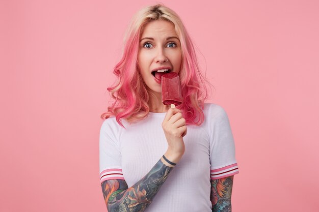Photo of beautiful girl with pink hair and tattooed hands, wearing a white t-shirt, looking and trying to bite off the ice cream, standing. Free Photo