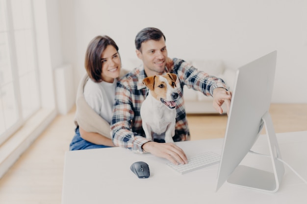 Photo of family couple make shopping on favourite website, enjoy time together, funny dog focused in monitor of computer, sits in coworking space. Premium Photo