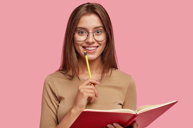 Photo of happy brunette woman with positive smile, carries textbook, holds pencil for writing Free Photo