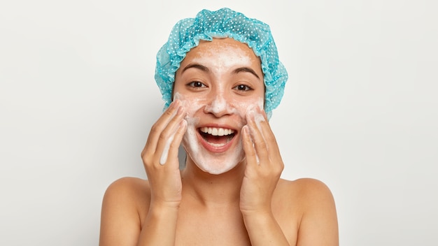 Photo of lovely female model with happy expression, washes face with foaming cleanser, wears wateproof showercap, pampers skin, stands shirtless, looks straightly. facial treatment Free Photo