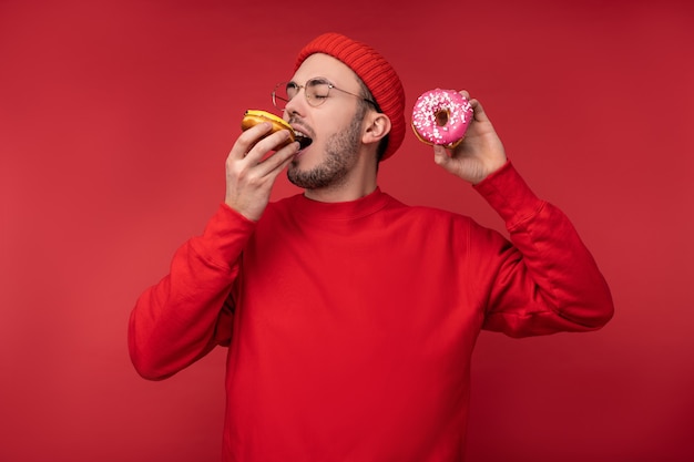 眼鏡と赤い服を着たひげを持つ幸せな男の写真 赤い背景の上に隔離されたお菓子ドーナツを食べることをお楽しみください プレミアム写真