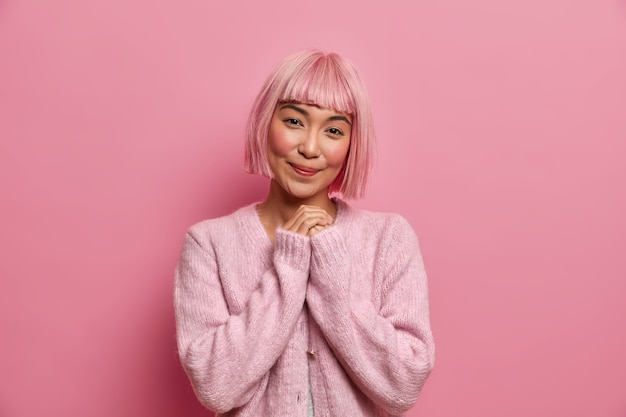 Free Photo Photo Of Pretty Asian Woman In Good Mood Keeps Hands Together Has Charming Smiling Expression Thinks About Something Pleasant Wears Pink Jumper Stands Indoor