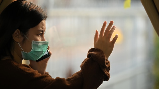 Photo of young woman wearing on medical mask using a smartphone while standing at the modern living room windows. woman keeping a quarantine herself at home. social distancing concept. Premium Photo