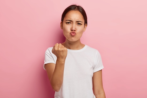 Picture of angry dissatisfied asian woman clenches fist with displeasure, keeps lips folded, makes outraged face expression, wears white clothing, threatens you, isolated on pink wall. Free Photo