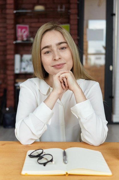 Free Photo Picture Of Attractive Successful Young European Female Writer With Blonde Hair Having Coffee At Cafe Sitting Alone At Wooden Table With Mug And Open Copybook Waiting For Friend For