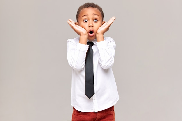 Picture of emotional funny surprised african american schoolboy in shirt and tie holding hands at his face, widening eyes and opening mouth widely, being shocked with astonishing unexpected news Free Photo