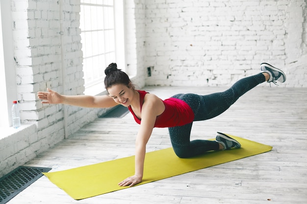 Picture of happy charming young female with hair bun doing physical training in light room, performing plank, raising opposite leg and arm, preparing body for summer. people and active lifestyle Free Photo