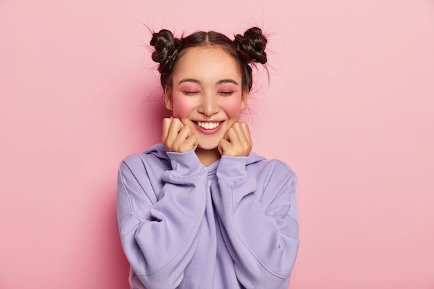 Pinup japanese woman with two combed hair buns touches cheeks, has smooth skin, wears vivid rosy makeup, piercing in nose, wears sweatshirt, smiles positively, isolated over pink background. Free Photo