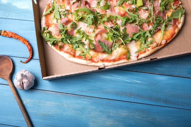 Pizza with bacon and arugula leaves in cardboard box with red chili and garlic bulb over wooden table Free Photo