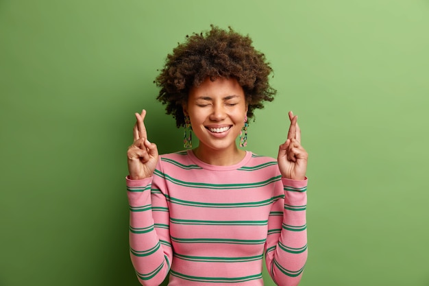Please i want it. cheerful young afro american woman smiles broadly dressed in casual striped jumper crosses fingers makes wish begs god for dream come true isolated over vivid green wall Free Photo