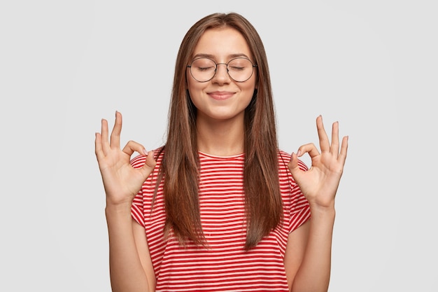 Pleased satisfied young female model makes zero gesture, wears transparent glasses, has long dark hair Free Photo