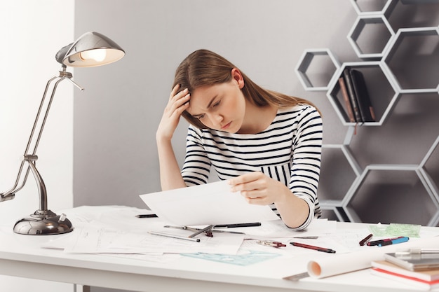 Portrait of beautiful young unhappy female engineer sitting at white table in cosy coworking space, looking at papers with upset expression being sad after finding mistake in blueprints. Free Photo