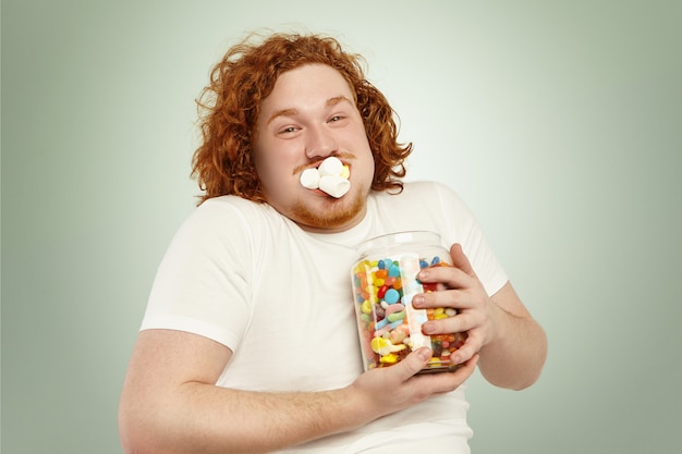 Portrait of cheerful young european overweight obese male with curly ginger hair consuming junk good, stuffing marshmallow into mouth and holding tight large glass jar, full of colorful sweets Free Photo