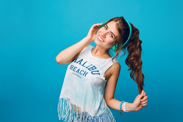 Free Photo Portrait Of Cute Girl With Long Curly Hair In Tail On Blue Background She Wears White T Shirt Shorts And Listening To Music With Blue Headphones