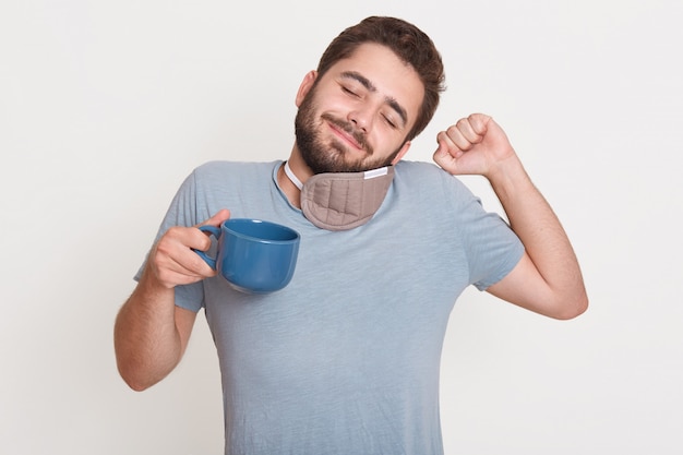 Portrait of delighted relaxed handsome young man closing eyes, waking up, having sleeping mask around neck, raising one arm, having fist Free Photo