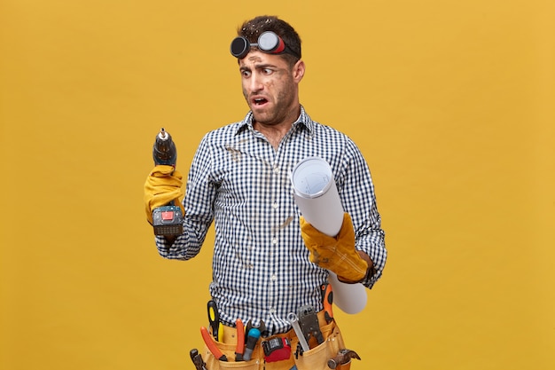 Portrait of dirty repairman having tool belt holding blueprint and drill machine looking at it with anger as it works badly. craftsman expressing his dissatisfaction with tools and instruments Free Photo