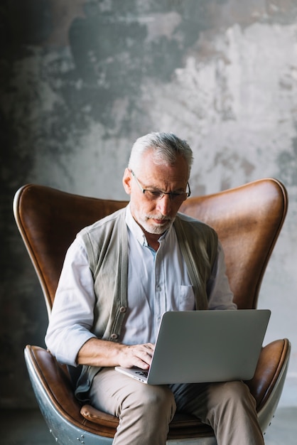 Portrait of elderly man sitting on chair using laptop ...