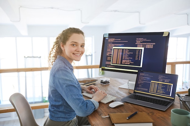 Portrait of female it developer smiling at camera while typing on keyboard with black and orange programming code on computer screen and laptop in contemporary office interior, copy space Premium Photo