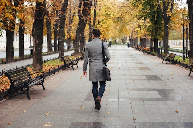 Portrait from back of young stylish guy in coat with bag strolling in city park, looking on colorful trees Free Photo