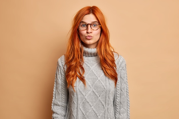 Portrait of good looking redhead woman keeps lips rounded has romantic face expression wants to kiss someone wears grey sweater. Free Photo