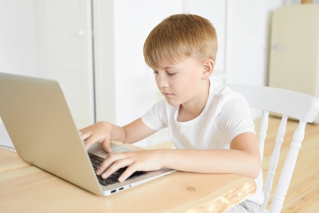 Portrait of handsome serious caucasian boy of school age sitting at wooden table using laptop computer, keeping hands on keyboard. education, leisure, people and modern electronic gadgets concept Free Photo