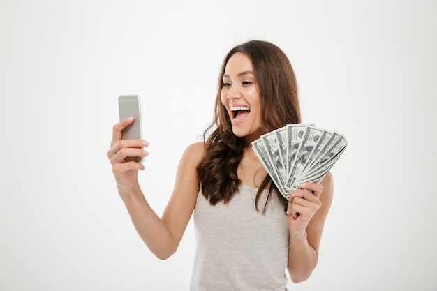 Portrait of happy joyous female 30s demonstrating lots of money dollar currency while using her mobile phone, isolated over white Free Photo