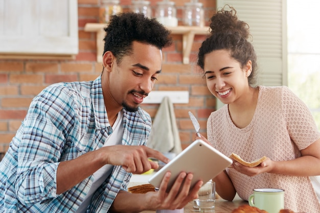 Portrait of mixed race hipster guy shows something at tablet computer to his wife who is busy making sandwiches 