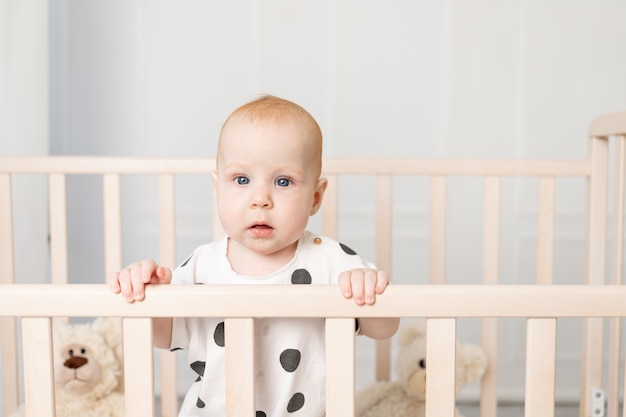 寝て カメラ テキストのための場所を見た後 明るい子供部屋のパジャマのおもちゃでベビーベッドに立っている8ヶ月の赤ちゃんの肖像画 プレミアム写真