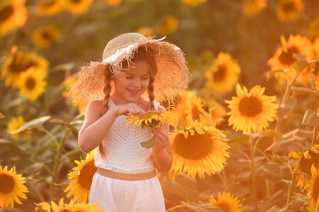 ひまわり畑で夕暮れ時の軽い服と大きな麦わら帽子でかわいい笑っている女の子の肖像画 彼女は手に大きな花を持っています プレミアム写真