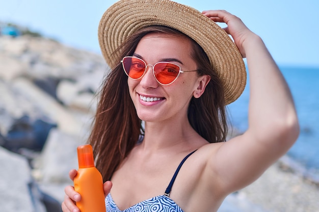 夏の晴れた日の海で日光浴中に水着 麦わら帽子 日焼け止めクリームのボトルと真っ赤なサングラスで幸せな笑顔の女性の肖像画 プレミアム写真