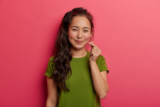 愛らしいブルネットの少女の肖像画は愛と幸福を広め ハートのサイン 指で愛情の韓国のシンボルを示し 明るいピンクの背景の上に分離された緑のtシャツを着ています 無料の写真