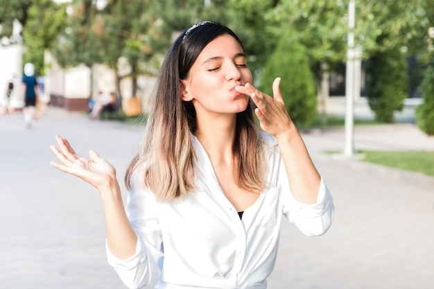 都市の公園で屋外の彼女の指をなめるとジャンクがおいしいファーストフードを楽しんでいる食べ物の完全な口で白いシャツと髪のベゼルで美しい女性の肖像画 プレミアム写真