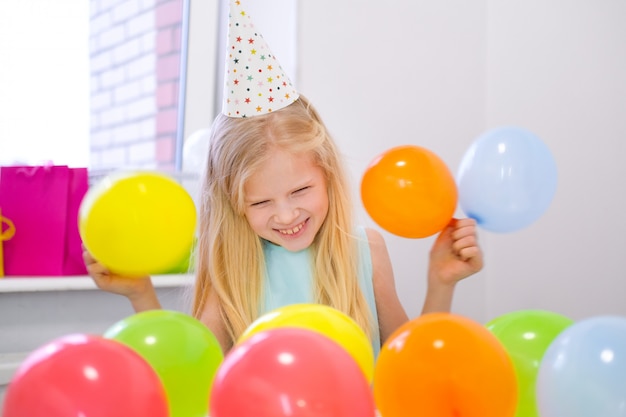 カメラの誕生日パーティーで笑っている金髪の白人少女の肖像画 風船でお祭りのカラフルな背景 縦写真 プレミアム写真