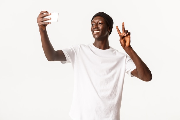 ビーニー帽の陽気なハンサムな黒人男性の肖像画 携帯電話で自分撮りを取り 幸せな 笑顔で平和のジェスチャーを示しています プレミアム写真