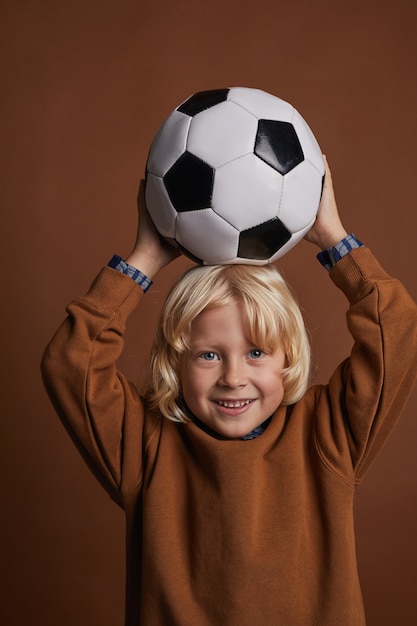 彼の頭の上にサッカーを保持し 茶色の背景を見て金髪のかわいい子供の肖像画 プレミアム写真