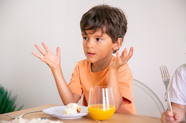 バースデーケーキを食べて オレンジジュースを飲むかわいい男の子の肖像画 食堂のテーブルに座って 手を上げて 目をそらしている愛らしい子供 子供の頃 お祝い 休日の概念 無料の写真